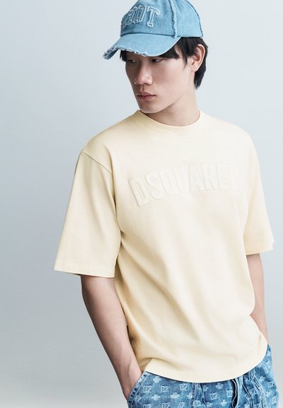 Jeune homme portant un t-shirt DSQUARED2 jaune pâle, un jean en denim bleu à motifs, et une casquette bleu clair usée, regardant vers la gauche.