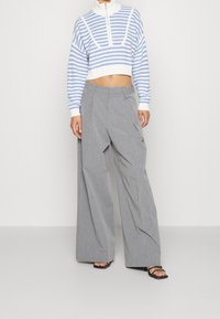 Donna che indossa un maglione corto a righe blu e bianche, pantaloni grigi a gamba larga e tacchi neri aperti davanti, in piedi contro uno sfondo semplice.