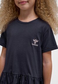 Dunkelblaues T-Shirt-Kleid aus weicher Baumwolle, mit kurzen Ärmeln, einem Rüschensockel und einem kleinen, pinkfarbenen bestickten Logo auf der Brust.