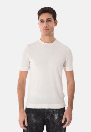 Uomo che indossa una maglietta a maniche corte bianca semplice in maglia e pantaloni neri con motivo macchiato bianco, in piedi su sfondo bianco.