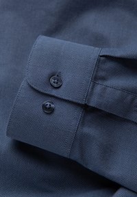 Tessuto di camicia blu scuro con una texture visibile e un polsino abbottonato, caratterizzato da due bottoni neri, ben piegato e esposto.