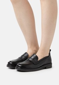 Black leather loafers with a smooth finish, featuring a stitched accent and a slight platform sole. Displayed on bare legs against a light background.