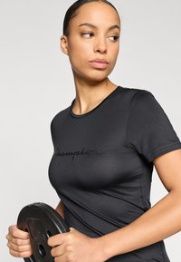 Μαύρο εφαρμοστό αθλητικό t-shirt με κοντά μανίκια και στρογγυλή λαιμόκοψη, με ανάγλυφο λογότυπο "Champion" στο στήθος. Κρα τάει μια δίσκο βάρους.