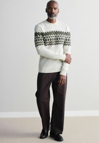 Helle Pullover mit grünem geometrischem Muster auf der Brust, kombiniert mit dunklen Hosen und schwarzen Schuhen. Einfaches, modernes Design.