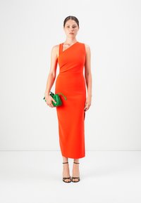 Frau trägt ein leuchtend orangefarbenes einseitiges Midikleid, hält eine kleine grüne Clutch und trägt schwarze Riemchenschuhe auf einem weißen Hintergrund.