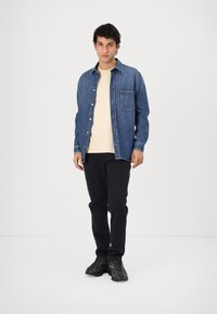Een blauwe denimshirt met een borstzak, gecombineerd met een lichtbeige T-shirt en een donkere broek. Het schoeisel heeft een gestructureerd zwart ontwerp.