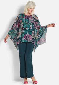 Doorzichtige bloemenblouse met belmouwen, voorzien van grote roze en groene bloemen op een donkere achtergrond, gecombineerd met teal broek en roze sandalen.
