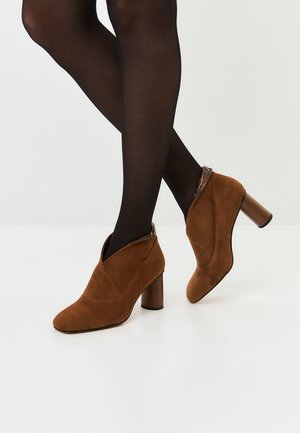 Jalat yang mengenakan legging hitam dengan sepatu bot pergelangan kaki suede coklat berhak blok dan trim bercorak ular di latar belakang putih.
