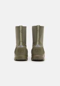 Botas de tobillo verde oliva con suelas de goma texturizadas, detalle de cremallera lateral y acentos acanalados. Presenta un material sintético suave y un logo.