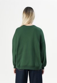 Persona con lunghi capelli biondi che indossa un ampio felpa verde scuro e jeans blu scuro, voltata di spalle contro uno sfondo bianco semplice.