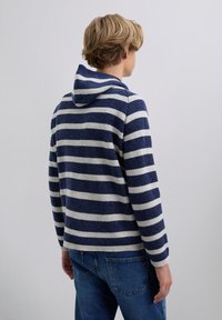 Henkilö, jolla on lyhyet vaaleat hiukset, pukeutuneena sinivalkoraidalliseen huppariin ja sinisiin farkkuihin, kääntynyt poispäin yksinkertaista taustaa vasten.