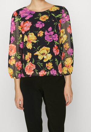 Blouse noire transparente à imprimé floral jaune, rose et orange, manches trois-quarts bouffantes, portée avec un pantalon noir sur une personne debout.