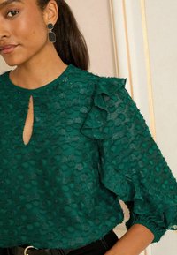 Blusa verde con texture e maniche a balze, dotata di scollatura a chiave e intricate fantasie a pois. Tessuto elegante con una leggera lucentezza.
