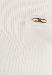 Weißes, geripptes Stoffmaterial mit einer kleinen rechteckigen Tasche, die mit einem goldenen, dekorativen Verschluss mit Kettendetail versehen ist.