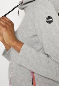 Haut à capuche zippé gris avec un tissu texturé, accent de fermeture éclair rose, logo noir sur la manche et capuche avec cordon de serrage. Gros plan sur des mains qui ajustent.