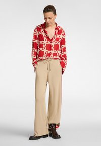 Camicia di seta rossa con motivo a catena beige, pantaloni beige a gamba larga con coulisse e strisce laterali, e stivaletti marroni con suola spessa.