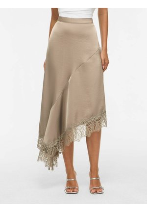 Kvinde iført beige asymmetrisk midinederdel med blondekant, hvid top og sølvfarvede hælsandaler, står mod en ensfarvet baggrund.