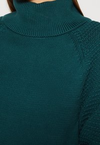 Pull à col roulé en tricot teal avec des accents texturés sur les épaules et un col côtelé. Matière douce d'épaisseur moyenne et coupe ample.