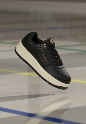 Melna ādas sporta apavi ar baltu zoli un bronzas krāsas Champion logotipiem, pacelti virs sporta zāles grīdas ar krāsainām laukuma līnijām.