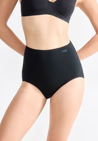 Donna che indossa un perizoma nero a vita alta e un reggiseno nero abbinato, in piedi contro uno sfondo chiaro e semplice.