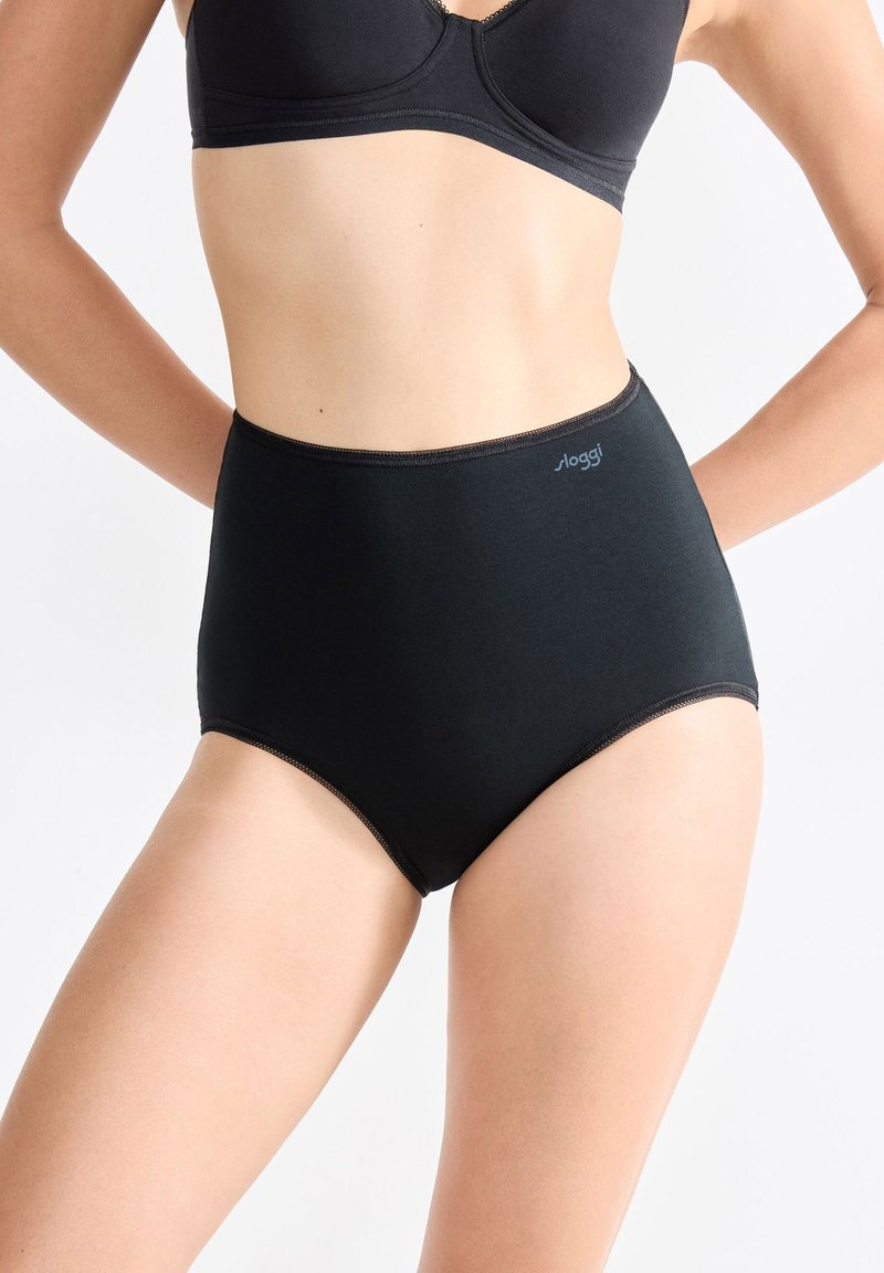 Donna che indossa un perizoma nero a vita alta e un reggiseno nero abbinato, in piedi contro uno sfondo chiaro e semplice.