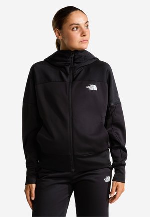 Kvinde iført en sort hoodie med lynlås og matchende bukser, begge med The North Face hvide logo, kigger til siden mod en hvid baggrund.