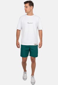Camicetta bianca a maniche corte con logo nero, abbinata a pantaloni corti verdi. Il modello indossa sneakers bianche con accenti rossi. Tessuto morbido e vestibilità casual.