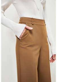 Pantalon marron taille haute à jambes larges, avec une texture lisse et des poches latérales. Associé à un haut blanc à manches longues côtelé.