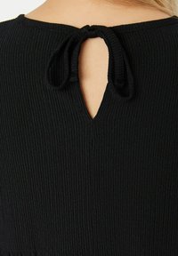 Top in tessuto a coste nere con scollo rotondo e dettaglio a chiave sulla schiena, fissato con un laccio sul retro. La texture è morbida e traspirante.