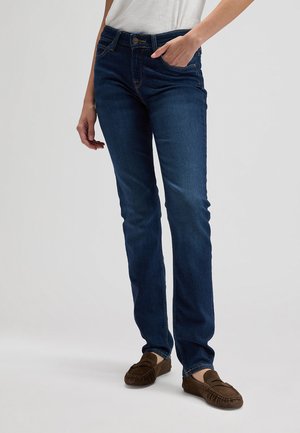 Osoba koja nosi tamnoplave traperice ravnih nogavica i smeđe cipele u stilu mokasina, s jednom rukom u džepu.