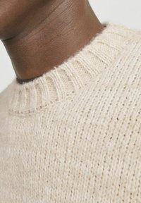 Beige knitted sweater with a ribbed crew neckline, featuring a textured pattern and a slightly fuzzy finish. Close-up view of the neck area.