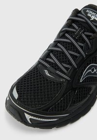 Sort atletiksko med mesh-overdel, reflekterende accenter og en struktureret sål. Snørebånd i sort med hvid syning. Buede designelementer.