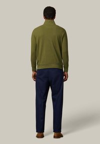 Suéter verde de punto con cuello alto; combinado con pantalones azul marino. Presenta puños y dobladillo acanalados, visto desde atrás. Calzado marrón visible.