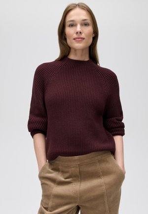 Vrouw draagt een donkerpaarse gebreide trui met opgerolde mouwen en lichtbruine ribfluwelen broek met handen in de zakken.