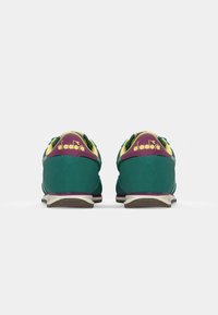 Groene suède sport schoenen met gele accenten, paarse hielcontrasten en witte zolen, voorzien van een logo aan de achterkant en een gestructureerd oppervlak.