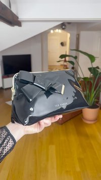 Clutch em pele sintética preta com um laço proeminente, detalhes em dourado e fecho de correr. Logótipo Ted Baker exibido na frente.