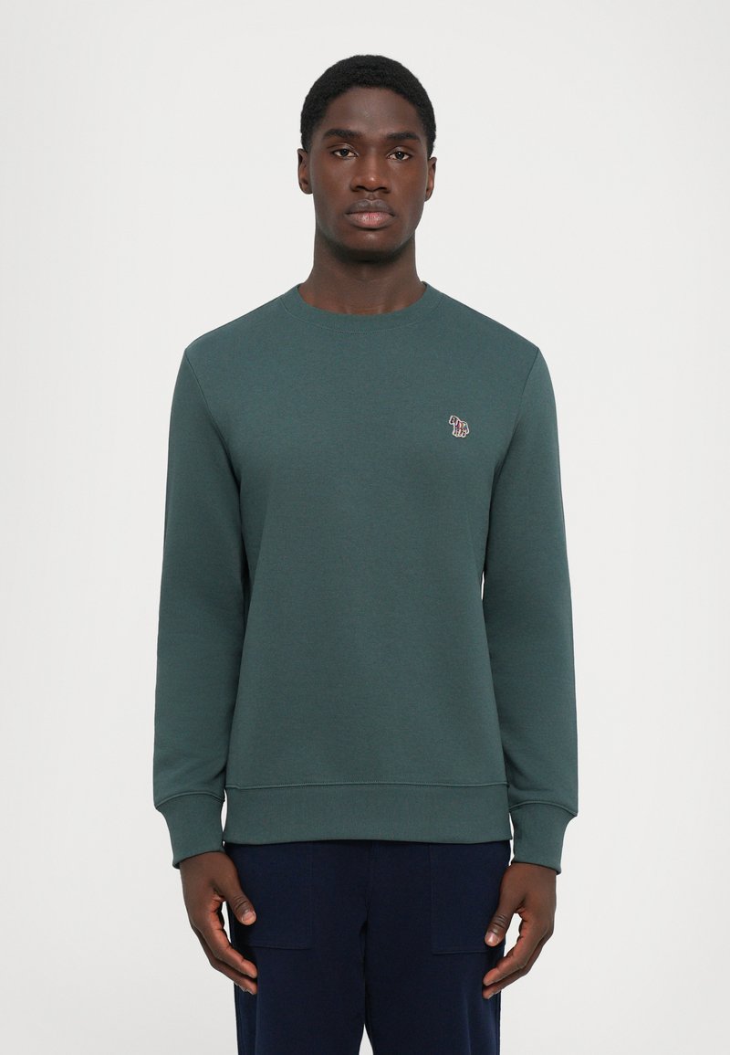 Joven vestido con un suéter de cuello redondo de color verde oscuro que tiene un pequeño logotipo de cebra multicolor en el pecho, de pie contra un fondo liso.