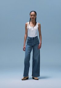 Weißes geripptes Top mit Knopfdetail, kombiniert mit hoch taillierten blauen Denim-Jeans im weit geschnittenen Stil und cremefarbenen spitzen Pumps.