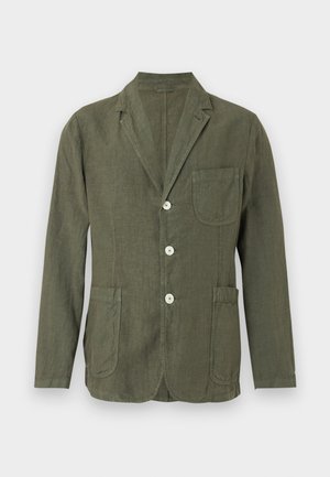 Man with curly hair wearing an olive green blazer, white shirt, beige wide pants, and black shoes, standing with hands in pockets.