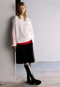 Weißes Langarm-Sweatshirt mit roten Grafiken, über einem roten Oberteil getragen, kombiniert mit einem schwarzen Rock und kniehohen schwarzen Stiefeln.