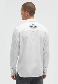 Weißes Langarmhemd mit einem Logo auf der Rückseite, Knopfmanschetten mit blauen Akzenten und einer lässigen Passform. Glatte Textur und entspanntes Design.