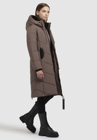 Brauner Puffer-Mantel mit Kapuze und schwarzen Akzenten, mit einem Chevron-Steppmuster, zwei seitlichen Taschen und einem durchgehenden Reißverschluss vorne.