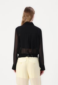 Blusa nera trasparente con colletto e vita elasticizzata, indossata sopra pantaloni giallo chiaro semi-trasparenti con tasche posteriori.