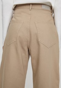 Pantalones de pierna ancha en beige, hechos de una tela gruesa, que presentan dos bolsillos traseros con forma de diamante, una cintura alta y un cinturón resistente.