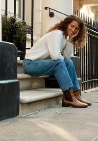 Bottines en cuir marron avec panneaux latéraux élastiques, posées sur une marche en béton. Le mannequin porte un pull crème et un jean bleu. Lumière naturelle.