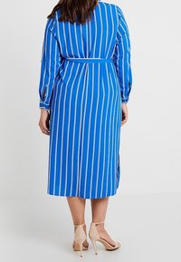 Donna che indossa un vestito midi blu con righe verticali bianche e una cintura in vita, abbinato a sandali con tacco alto e cinturini alla caviglia beige, mostrata di spalle.