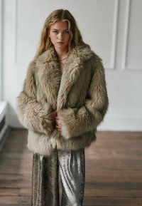 Faux fur frakke i lys brun med en fyldig tekstur, bred krave og lange ærmer. Matchende med skinnende sølvfarvede vide bukser.