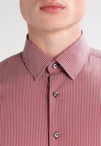 Chemise à rayures rouges avec un col boutonné, des boutons gris et une texture de tissu lisse. Rayures verticales dans différentes nuances de rouge.