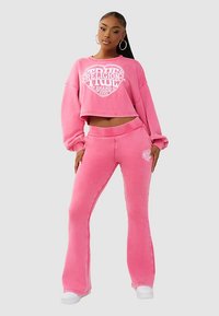 Roze cropped sweatshirt en bijpassende uitlopende broek van zachte stof, met een witte logo-ontwerp in het midden. Witte sneakers maken de outfit compleet.