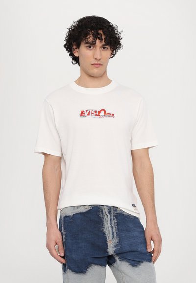 T-shirt en coton blanc à manches courtes, avec un logo graphique rouge et bleu sur le devant. Porté avec un short en denim usé.